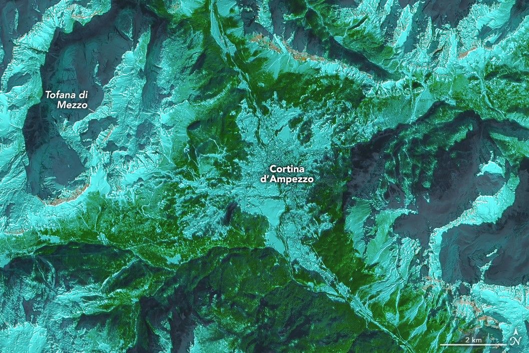 A false-color satellite image shows the town of Cortina d’Ampezzo, Italy, surrounded by tall mountains in the Dolomites. Snow appears light blue, forested areas are green, and bare cliff bands and light brown.